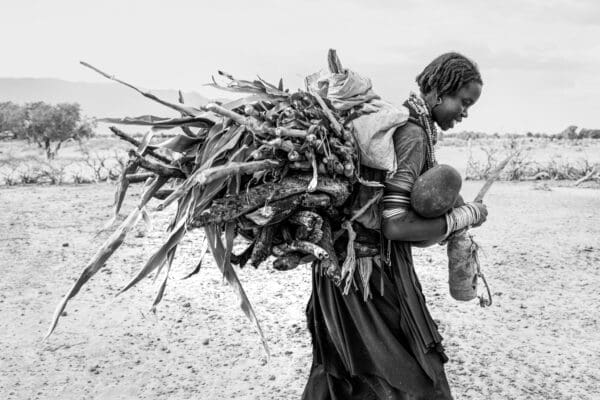 A woman from the Abore tribe walks while holding her young toddler in her arms. In one hand she holds the machete she used to cut the sorghum she carries in a bundle on her back. In the bundle is also a large stack of wood, most that will most likely be used to cook with. It's possible she will use the wood to cook the sorghum.
