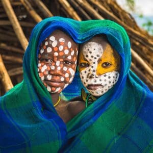 Two boys have their faces painted and are framed by a blue and green blanket