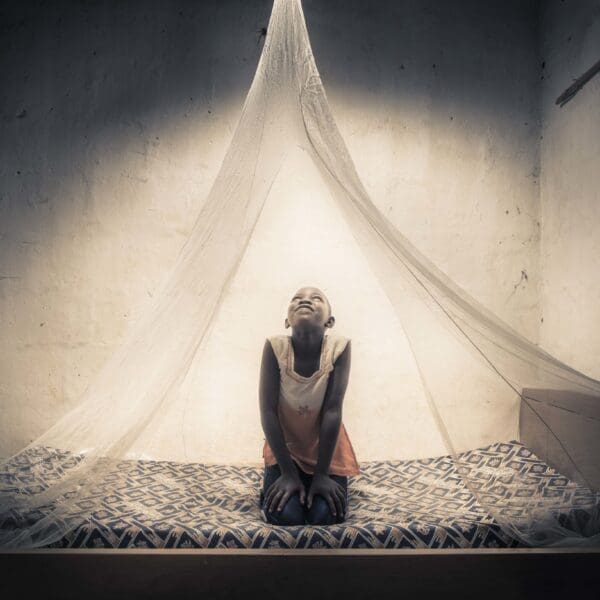 A girl sits on a bed smiling with a mosquito net around her.