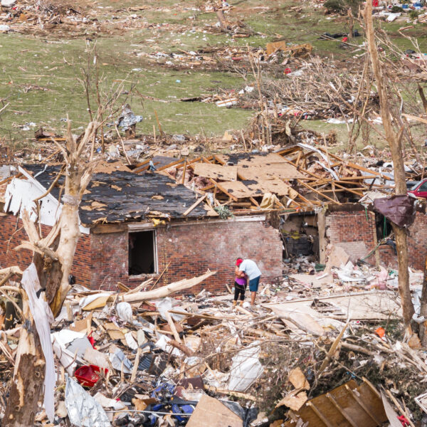 Two people embrace in the wreckage of a tornado-damaged home in London, Kentucky, surrounded by debris and broken trees.